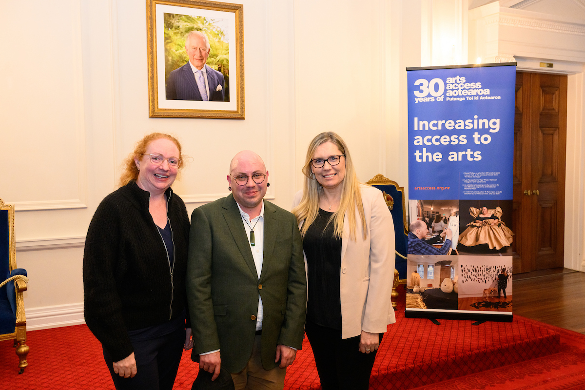 Theresa Cooper, Stace Robertson and Rahera Turner at the Government House High Tea event. They are standing to the left of the Arts Access Aotearoa 30-year banner and in front of a portrait of King Charles. 